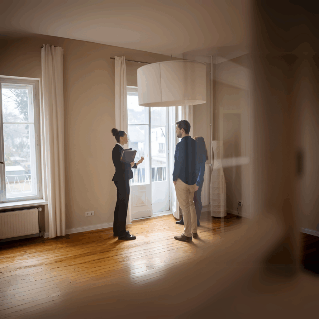 Immobilienmaklerin zeigt einem Paar eine helle, leere Wohnung mit Holzboden und großen Fenstern bei einer Besichtigung.